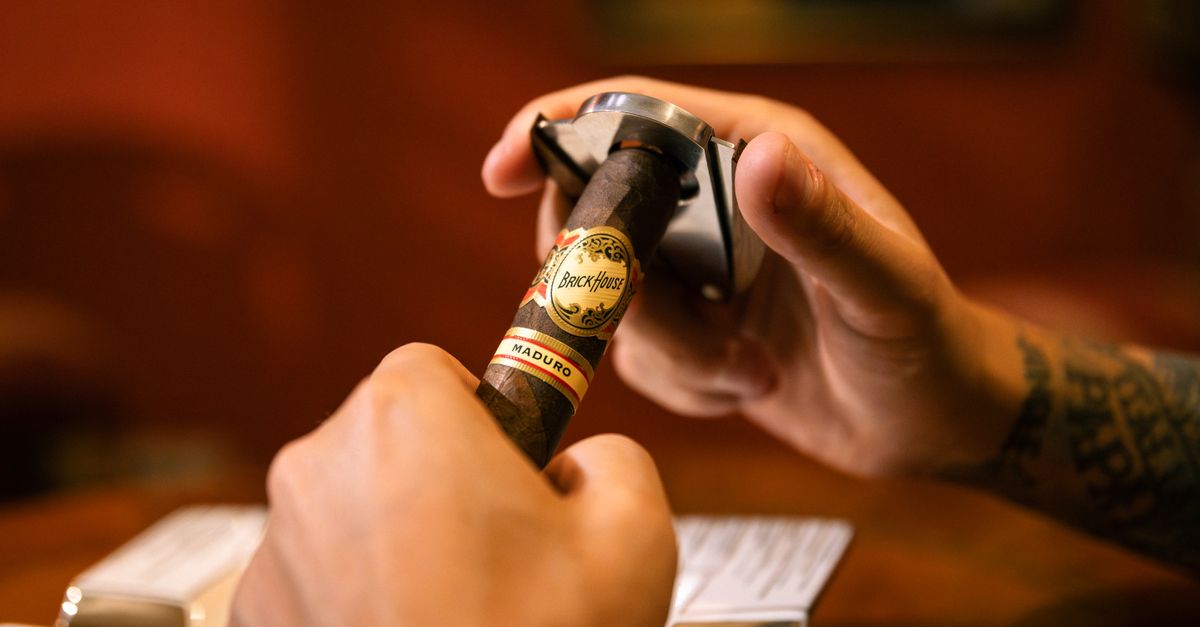 A close-up of hands using a cigar cutter on a BrickHouse Maduro cigar indoors.