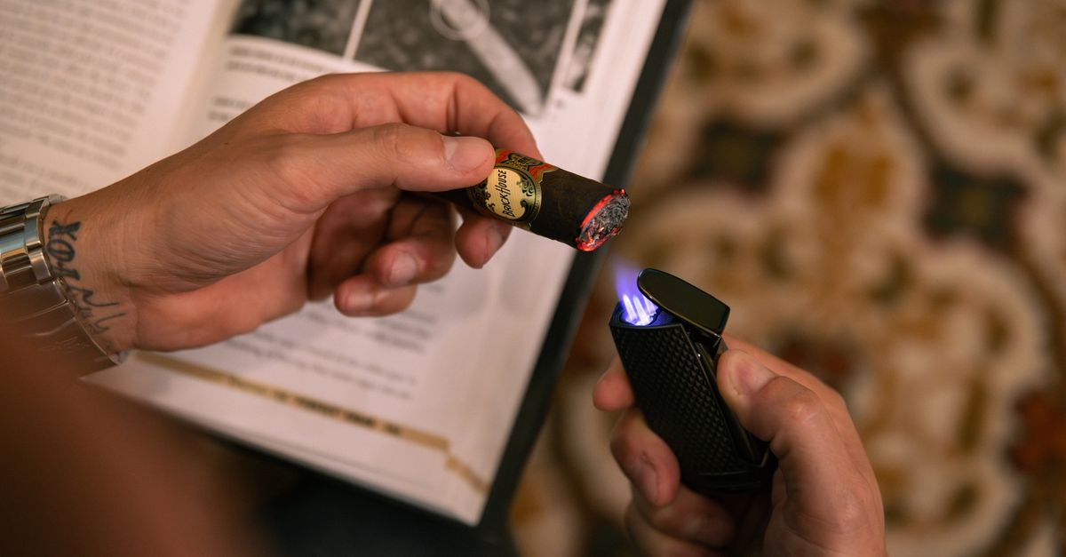 Close-up of hands lighting a cigar with a sleek lighter over an open book on a patterned rug.