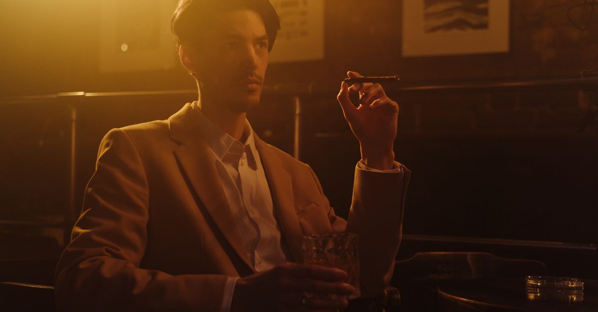 A man in a suit relaxes with a cigar and whiskey in an elegant setting.