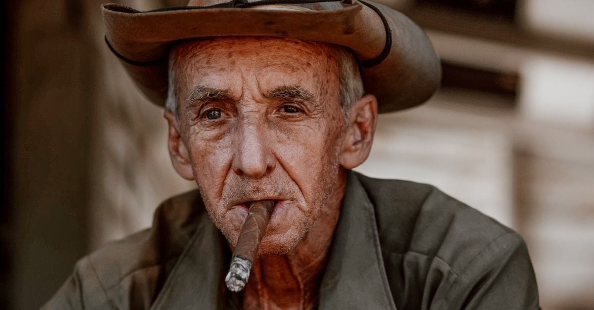 Serious aged male in cowboy hat sitting on street with blurred background while smoking cigar and looking away thoughtfully in daylight